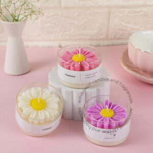 Decorative handcrafted Daisy candles in pink, purple, and white displayed in clear round containers on a pastel pink surface, with a white vase and pink teacup in the background.