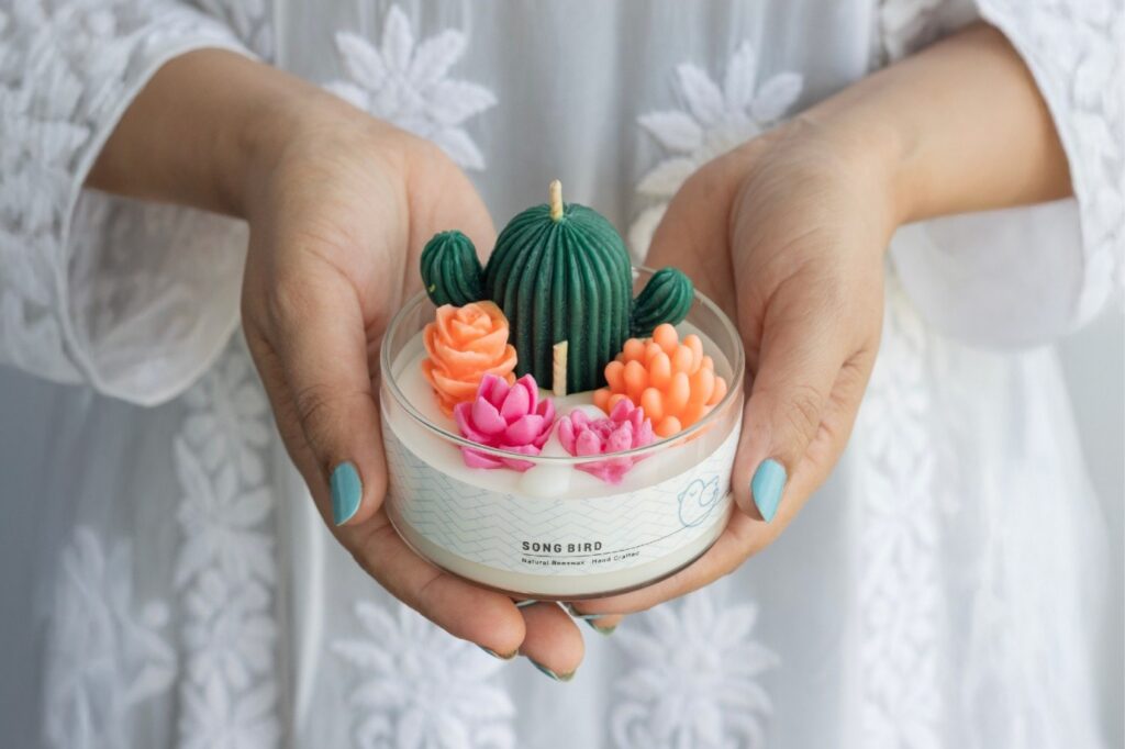 A Lady holding a beautifully crafted Song Bird candle featuring a green cactus and colorful floral designs, made with natural beeswax, perfect for affordable luxury birthday gifts.