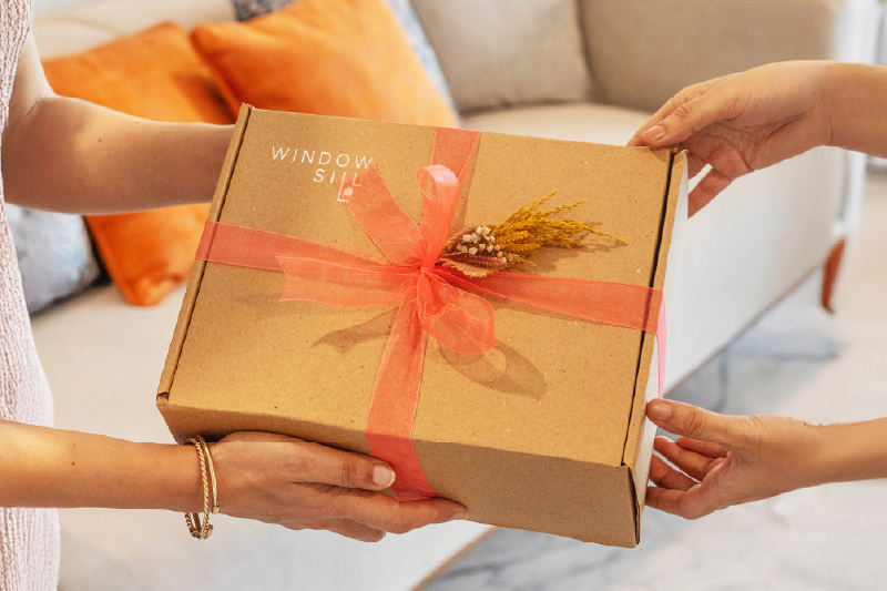 Hands exchanging a beautifully wrapped housewarming gift box with kraft paper, coral ribbon, and dried flowers, representing thoughtful unique housewarming gifts for welcoming friends to their new home.
