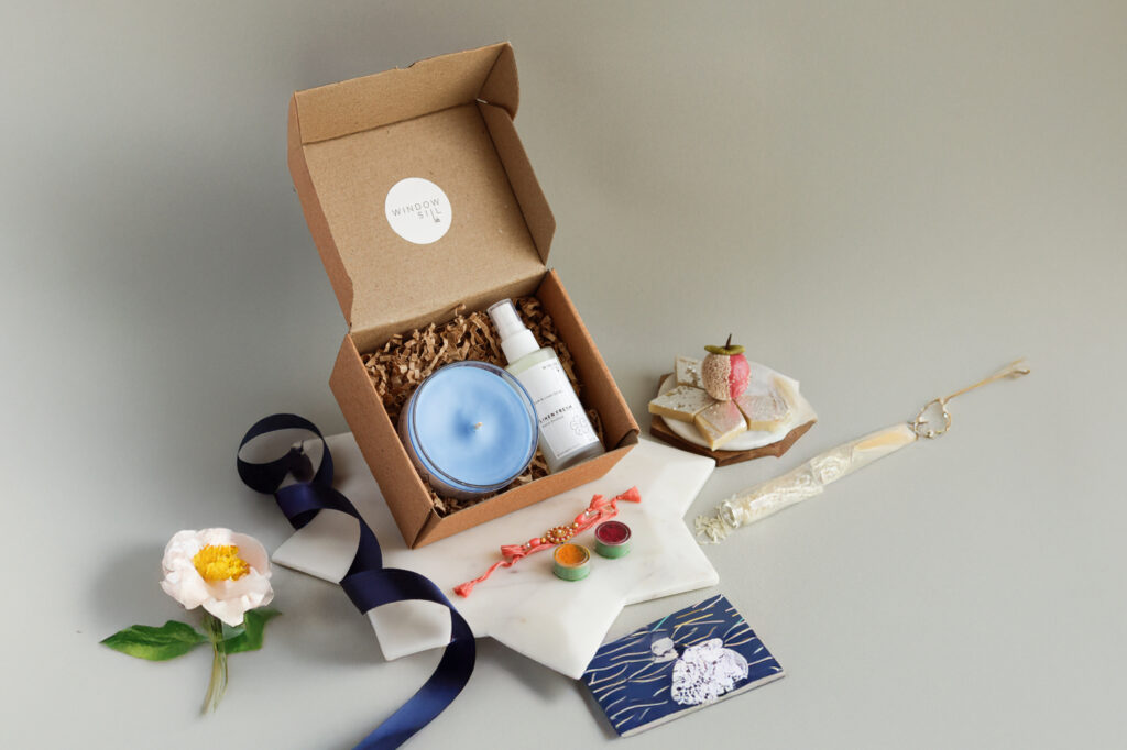 Windowsill Rakhi gift hamper with blue scented candle, wellness mist, roli-chawal set, and festive sweets.